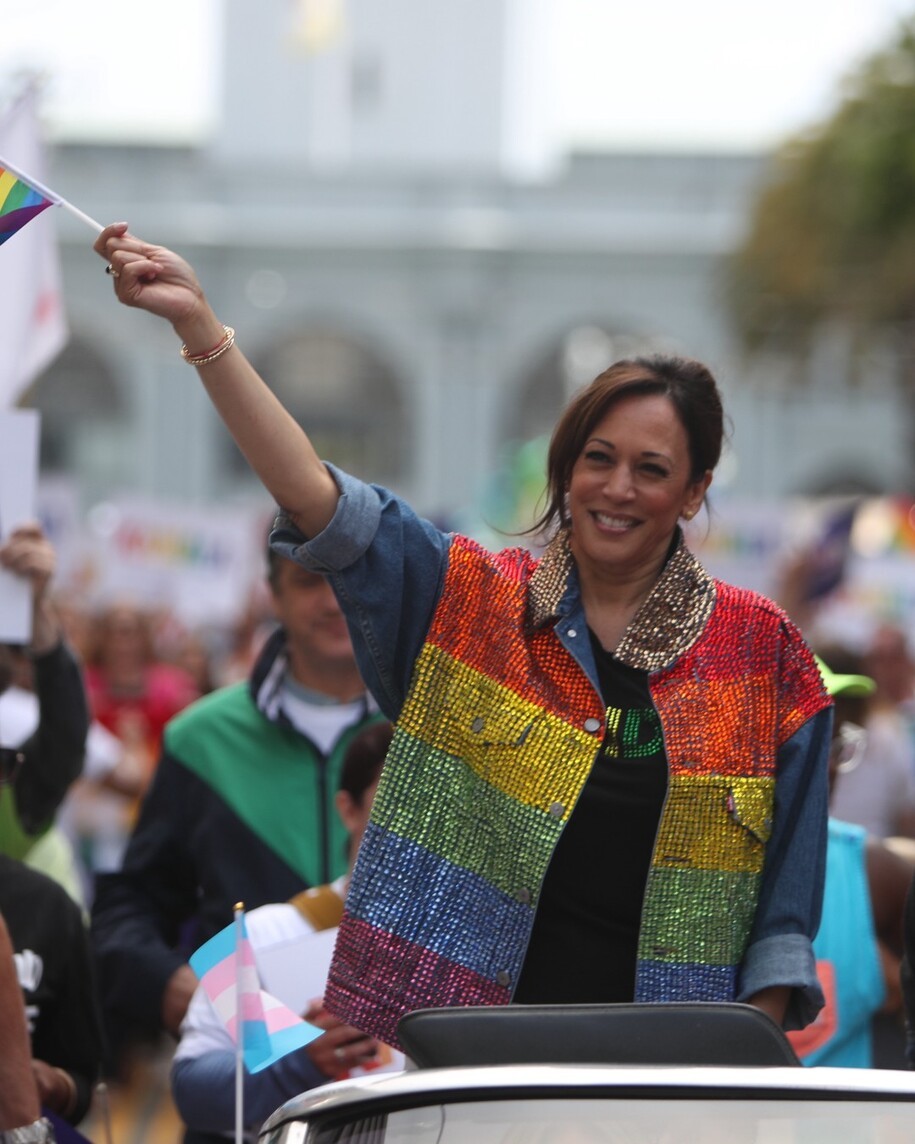 Vice President Kamala Harris waves a rainbow flag to a crowd from a convertible car at a Pride Parade, wearing a rainbow colored sequined jacket.