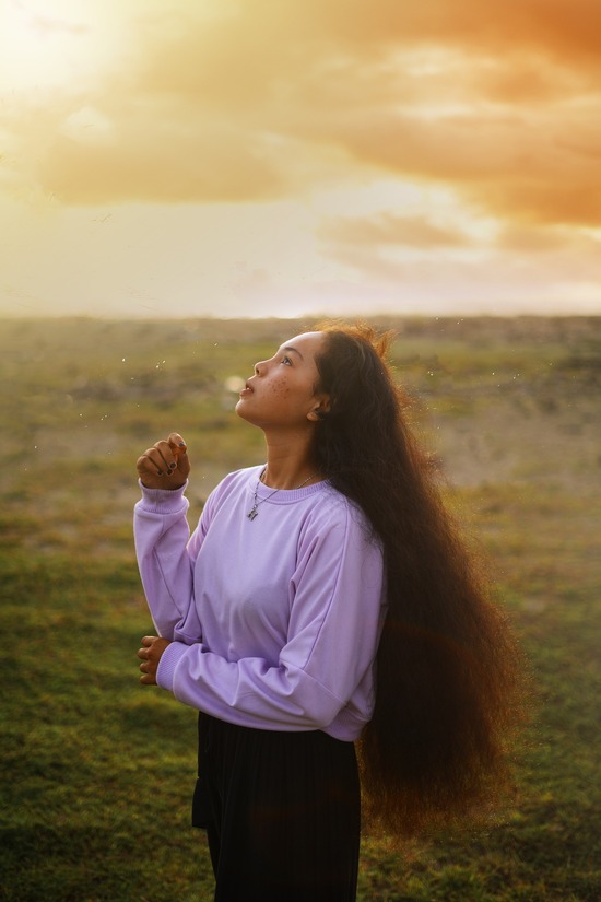 Asian or possibly Native American woman with very long thick red-brown hair gazes upward, lit by a sun behind clouds.
