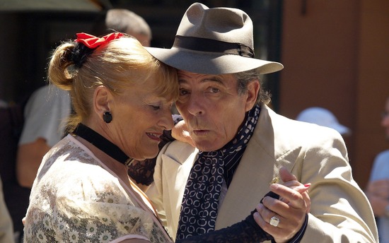 elderly couple dancing a tango. He is wearing a fedora, she has a red bow in her hair.